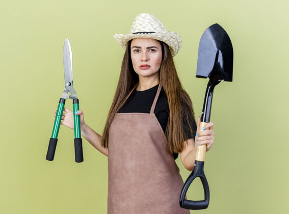 女孩自信美丽的园丁女孩穿着制服 戴着园艺帽 拿着剪子和铁锹 橄榄绿的背景上孤立帽子园艺制服