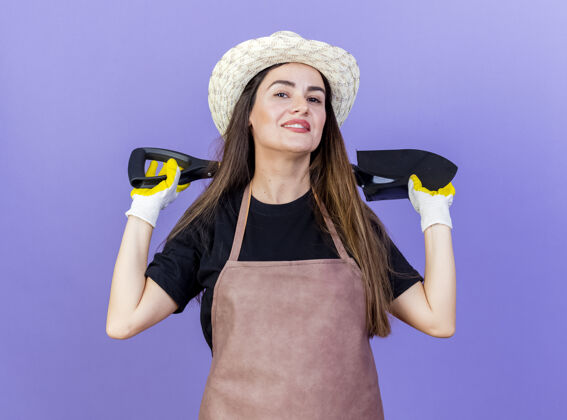微笑微笑美丽的园丁女孩穿着制服 戴着园艺帽和手套 脖子后面拿着铲子 蓝色背景上孤立着园艺黑桃花园