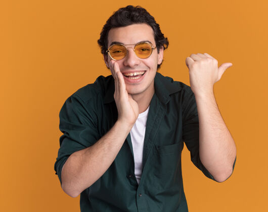 拇指身穿绿色衬衫 戴着眼镜的年轻人站在橙色的墙上 面带微笑 手靠近嘴边 拇指指向一边侧身年轻人手