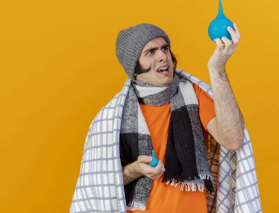 包裹关心的年轻病人戴着冬天的帽子 围巾裹着格子布 抬起头 看着隔离在橙色背景上的灌肠剂养育格子布疾病