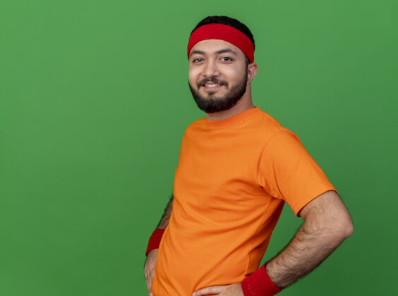 年轻微笑的年轻运动男子戴着头带和腕带把双手放在臀部隔离在绿色背景与复制空间腕带微笑臀部