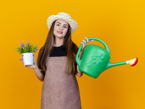 举行微笑美丽的园丁女孩穿着制服 戴着园艺帽 拿着浇水罐 花盆里的花被隔离在橙色的背景上花园花制服