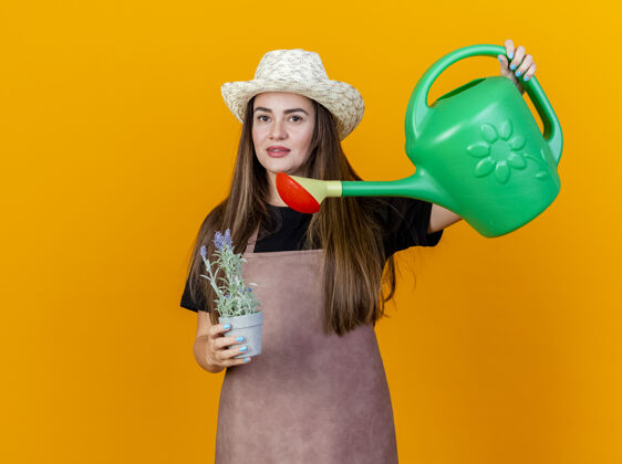 请很高兴美丽的园丁女孩穿着制服 戴着园艺帽 拿着花盆浇花 浇水罐隔离在橙色的背景上制服浇水帽子