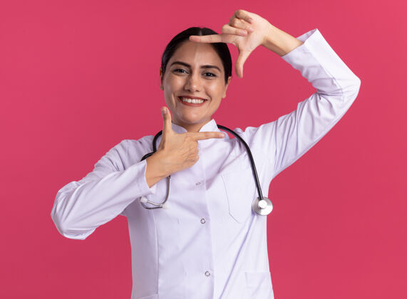 展示年轻的女医生 穿着医用外套 手持听诊器 面带微笑 站在粉红色的墙上 露出手指架着的样子外套框架微笑