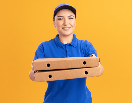 女人快乐的年轻送货员身穿蓝色制服 戴着帽子 手里拿着比萨饼盒 微笑着自信地站在橙色的墙上看着前面站着微笑自信