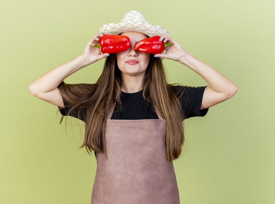 女孩一个穿着制服的漂亮园丁女孩戴着园艺帽 眼睛上拿着辣椒 橄榄绿的背景上孤立着帽子眼睛园艺
