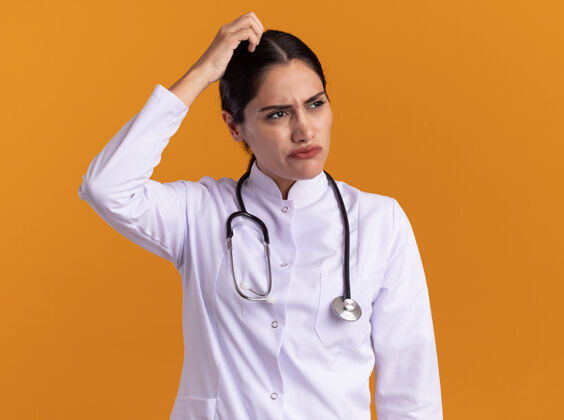 年轻年轻的女医生 穿着医用外套 脖子上戴着听诊器 困惑地看着一边 站在橙色的墙上挠头头站旁边