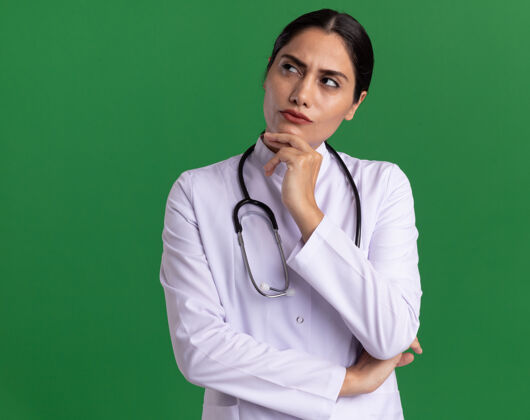 医生身穿医用外套 脖子上戴着听诊器的年轻女医生抬起头 脸上带着沉思的表情 想着站在绿色的墙上脖子表情脸