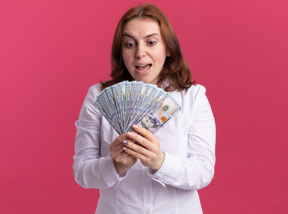 现金穿着白衬衫的年轻女子拿着现金看着钱 站在粉红色的墙上高兴又兴奋抱着女人兴奋
