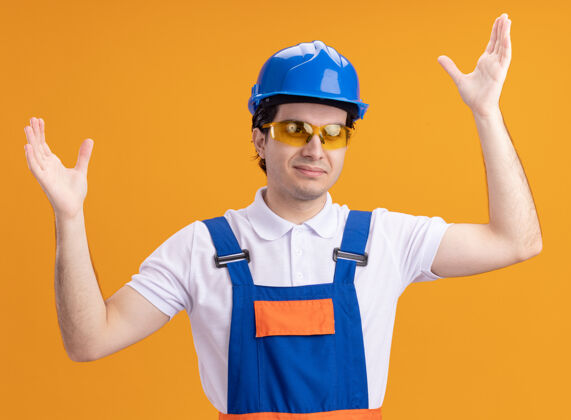 男人年轻的建筑工人身穿建筑制服 戴着安全帽 戴着眼镜 高举双臂站在橙色的墙上 愉快而积极地看着前方站立安全举起