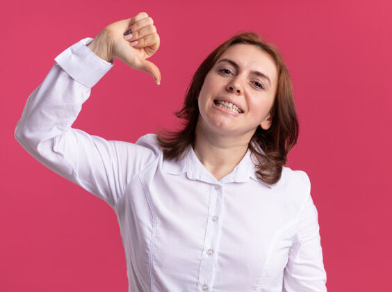 自信身穿白衬衫的年轻女子站在粉红色的墙上 自信地微笑着用拇指指着自己拇指她自己衬衫