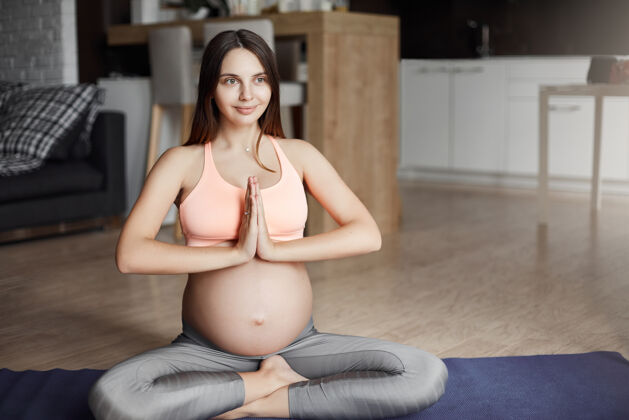 人穿着运动服的漂亮的怀孕白人女性 坐在滚动垫上做瑜伽姿势 双手合十 一边微笑一边放松 想着宝宝冥想身体医疗