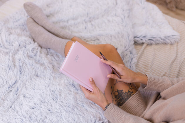 女穿着羊毛袜子和毛衣的女人 粉色笔记本上写着2021 臀部有大纹身女人坐在卧室的家里的床上卧室腿爱好