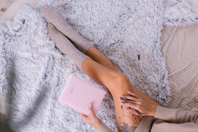 坐着穿着羊毛袜子和毛衣的女人 粉色笔记本上写着2021 臀部有大纹身女人坐在卧室的家里的床上休息计划者住宿