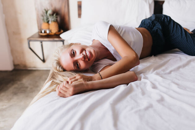 光疲惫的女人穿着深色裤子躺在床单上微笑着室内拍摄金发美女在床上摆姿势床金发女士