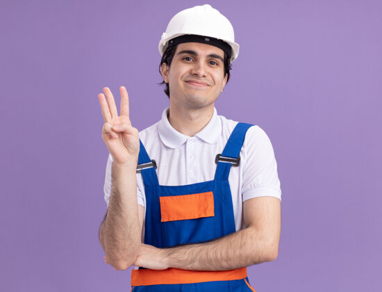 脸年轻的建筑工人穿着建筑制服 戴着安全帽 面带微笑地看着前面 三号站在紫色的墙上微笑男人头盔