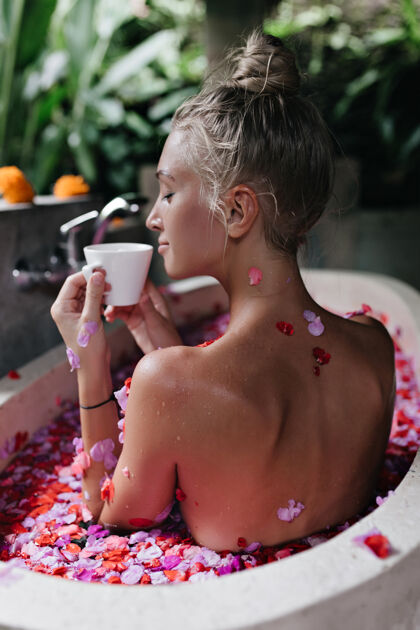 花瓣快乐的白人妇女坐在浴池里用玫瑰花瓣 闭着眼睛喝茶照片背面是一位受启发的白人女模特 她在早晨的温泉浴场里喝着咖啡咖啡人卫生