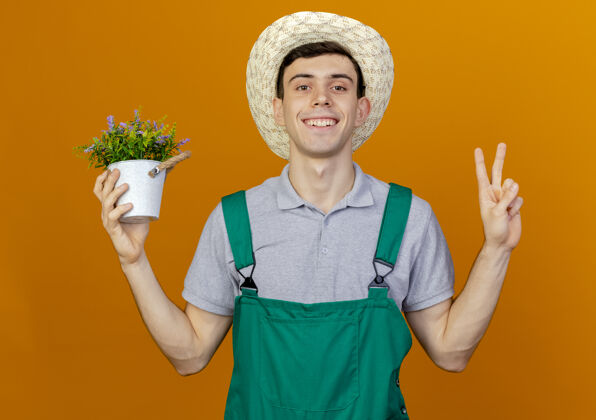 手势微笑的年轻男园丁戴着园艺帽 手持花盆 打着胜利手势年轻园艺花盆