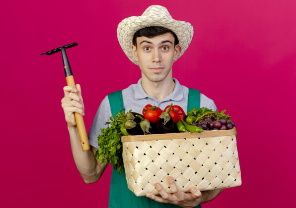 男性高兴的年轻男园丁戴着园艺帽拿着菜篮和耙子磨损帽子蔬菜