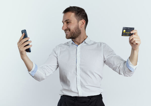 商人笑容可掬的帅哥拿着信用卡 看着隔离在白墙上的手机持有优雅男人