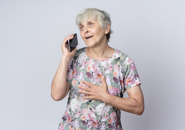 电话惊讶的老妇人把手放在下巴上拿着电话看着隔离在白墙上的一边惊喜老人壁板