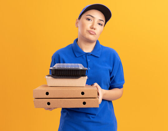 年轻身穿蓝色制服 头戴鸭舌帽 拿着比萨饼盒和食品包的年轻送货员站在橙色的墙上 脸上带着悲伤的表情看着前方表情制服食物