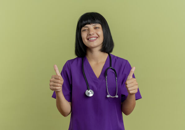 手微笑着的年轻黑发女医生穿着制服 手持听诊器 竖起大拇指 两手分开放在橄榄绿背景上 留有复印空间背景女人拇指