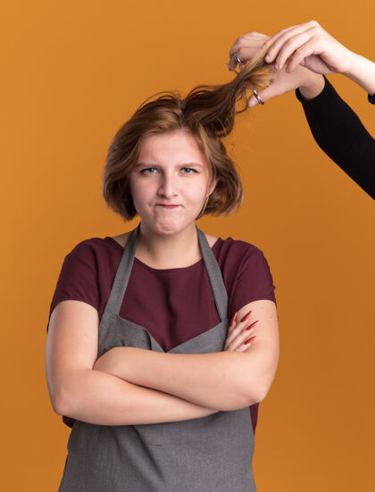 有人年轻漂亮的女理发师围着围裙看着前面不高兴和生气 因为有人站在橘色的墙上剪她的头发生气头发理发师