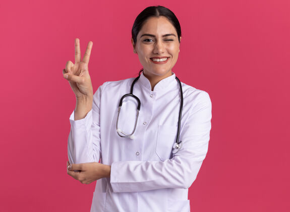 听诊器年轻的女医生穿着医用外套 手持听诊器 面带微笑 站在粉红色的墙上 脸上露出v字标志表演脸微笑