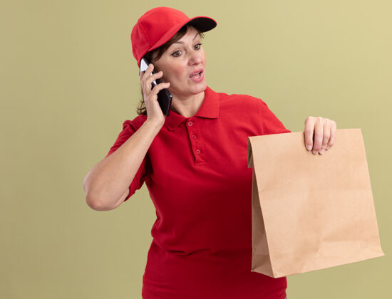 手机身穿红色制服 戴着帽子的中年送货妇女拿着纸包 站在绿色的墙壁上讲着手机 看上去很困惑送货包装站着