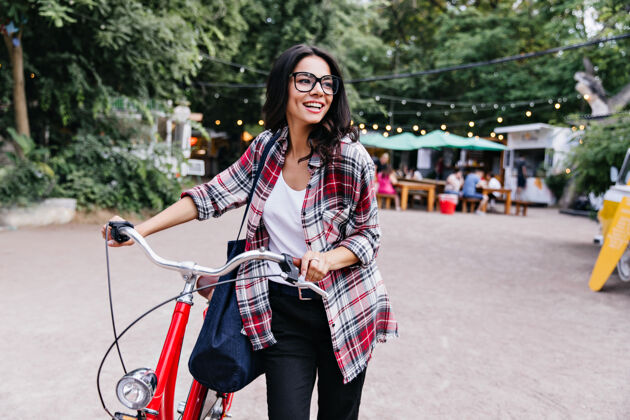 女人一位年轻漂亮的黑发女士骑着自行车站在街上一张有兴趣的黑发女孩穿着黑色裤子在周末玩的照片休息乐趣自行车
