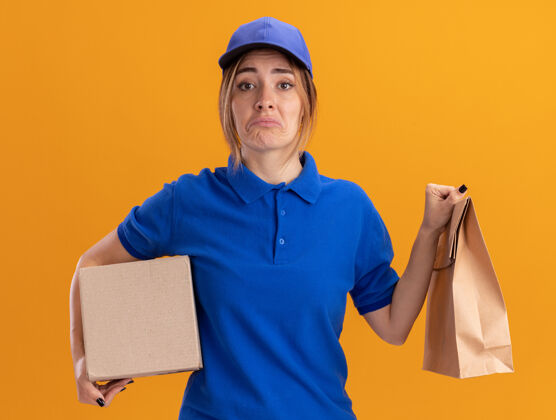 悲伤穿着制服的悲伤的年轻漂亮的送货女孩拿着橙色的纸包和名片盒包装漂亮年轻