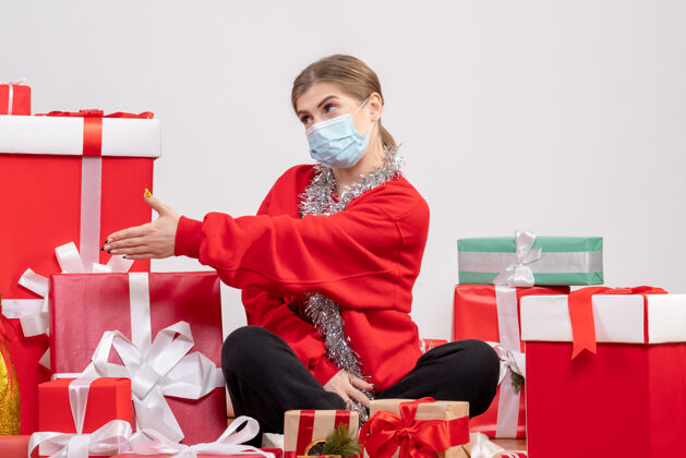 圣诞节正面图：年轻女性戴着无菌面具坐着 手里拿着圣诞礼物礼物地板坐