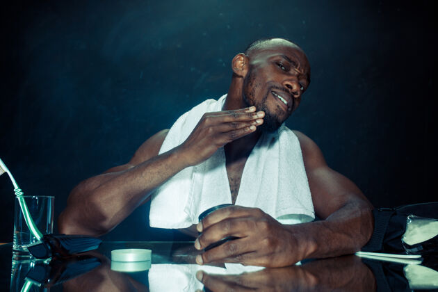 镜子在卧室里 一个年轻的非洲男人在家刮胡子后坐在镜子前房子奶油毛巾