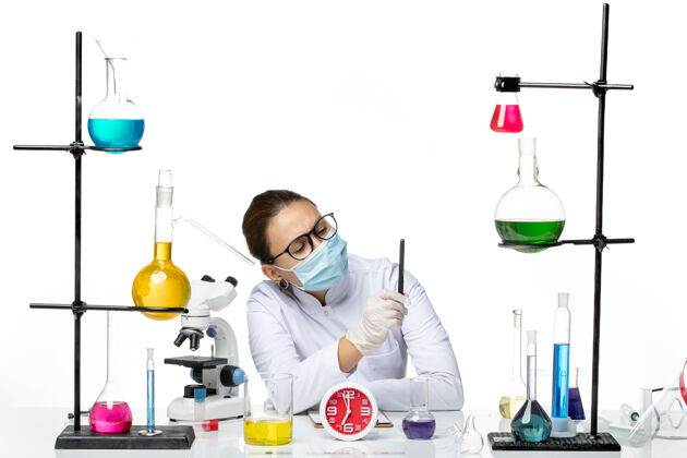 面罩前视图穿着医疗服的女化学家戴着面罩坐着拿着溶液拿着笔在白色背景上飞溅实验室病毒化学covid拿着药品钢笔