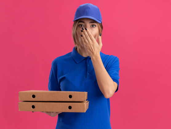 年轻一个穿着制服的年轻漂亮的送货女孩把手放在嘴上 手里拿着粉红色的比萨饼盒披萨手漂亮