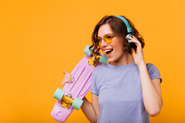 女性戴着黄色太阳镜的漂亮女孩戴着耳机听音乐穿着滑板唱着最喜欢的歌的时尚女模特的肖像酷放松无忧无虑