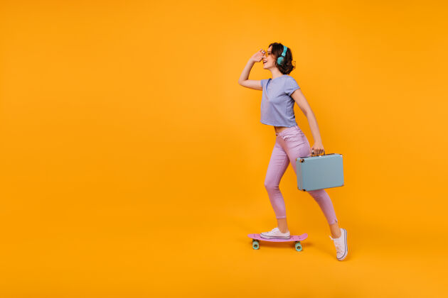 酷穿着粉色裤子的梦幻女孩站在滑板上听音乐灵感来源于戴着耳机的卷发女模特和蓝色的手提包合影音乐休闲女性