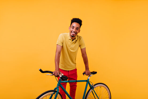 非洲感兴趣的非洲男模穿着红色裤子微笑着一幅充满灵感的黑人站在绿色自行车旁的画像休闲活跃美国