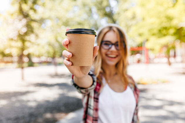 成人在公园里喝拿铁的漂亮女孩在笑前景是一杯咖啡的金发女人的模糊肖像娱乐肖像放松