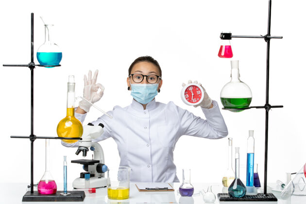 研究前视图：身穿医疗服的女化学家 戴着面具 白色背景上拿着时钟病毒实验室化学冠状病毒-飞溅医生专业医学