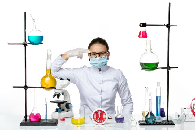 医生前视图：穿着医疗服的女化学家戴着面罩坐在白色办公桌上 带着溶液化学病毒实验室病毒-飞溅病毒医学视图