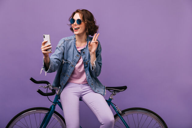 运动员穿着紫色裤子的迷人女孩在自拍穿着牛仔夹克的幽默小姐坐在自行车上配件波浪发卷发