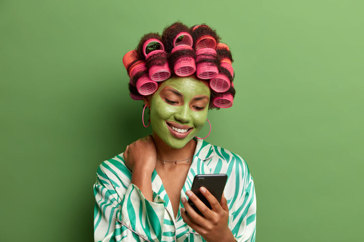 治疗喜气洋洋的女士专注于智能手机的展示 在阅读信息时积极微笑 穿着休闲服装 戴着美容面具 关心皮肤 卷发器打造完美发型家庭应用使用