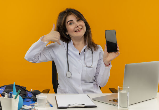 剪贴板快乐的中年女医生穿着医用长袍和听诊器坐在办公桌前用医疗工具剪贴板和笔记本电脑做通话手势显示手机被隔离电话表演手机