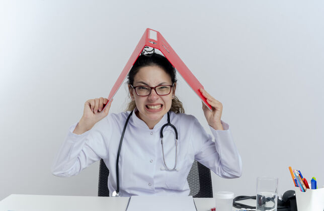 人压力重重的年轻女医生穿着医用长袍 戴着听诊器和眼镜坐在办公桌旁 手里拿着医疗工具 头上拿着文件夹抱着眼镜女士
