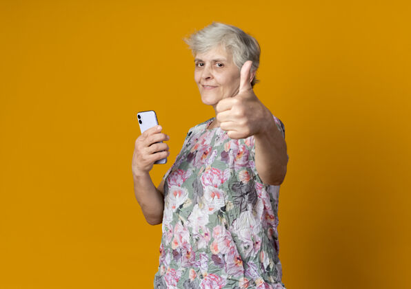 老人高兴的老妇人站在一边拿着电话 竖起大拇指孤立地站在橙色的墙上电话向上老人