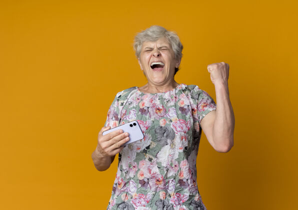向上快乐的老妇人拿着电话 举起拳头孤立地站在橙色的墙上老人举起快乐