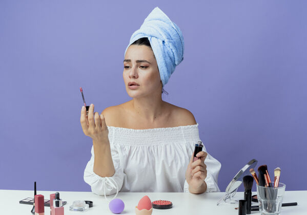 漂亮美丽的心烦意乱的女孩裹着毛巾坐在桌边 拿着化妆工具 看着隔离在紫色墙上的睫毛膏毛巾包女人