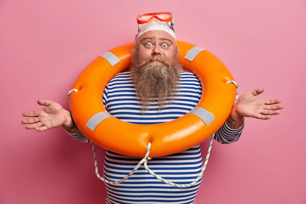 困惑犹豫不决的疑神疑鬼的胡子男人侧着手 感到困惑 戴着泳帽 护目镜和水手t恤 在粉红色的墙上与充气的救生圈隔离海滩上超重的救生员潜水人脂肪